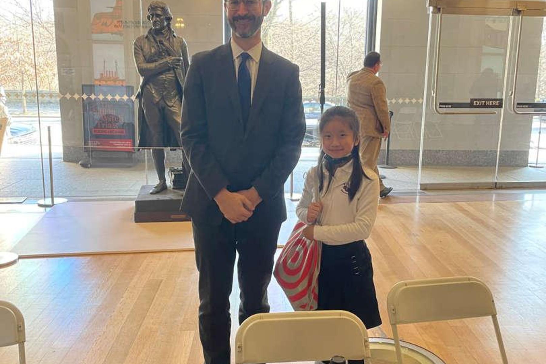 Man in a suit stands next to a girl with a striped bag at an event venue.