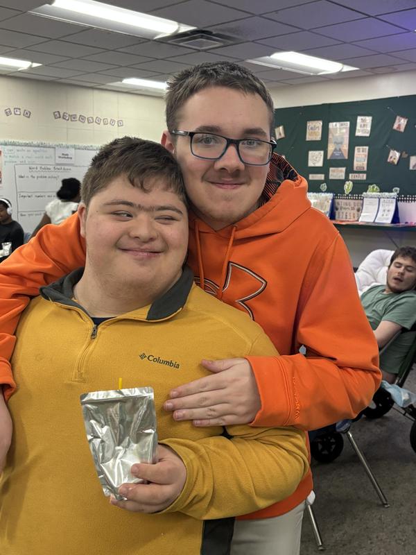 Two boys pose together smiling, one holding a juice pouch in a classroom.