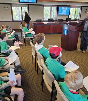 A group of children in green shirts listens to a speaker at a debate event in a conference room.
