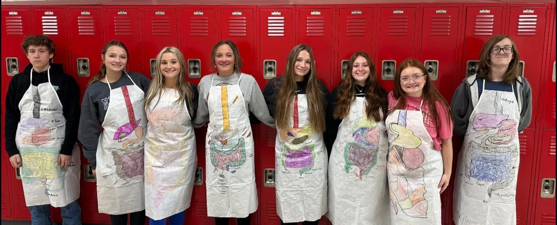 2024-25 Eight students in decorated Anatomy aprons stand against red lockers.