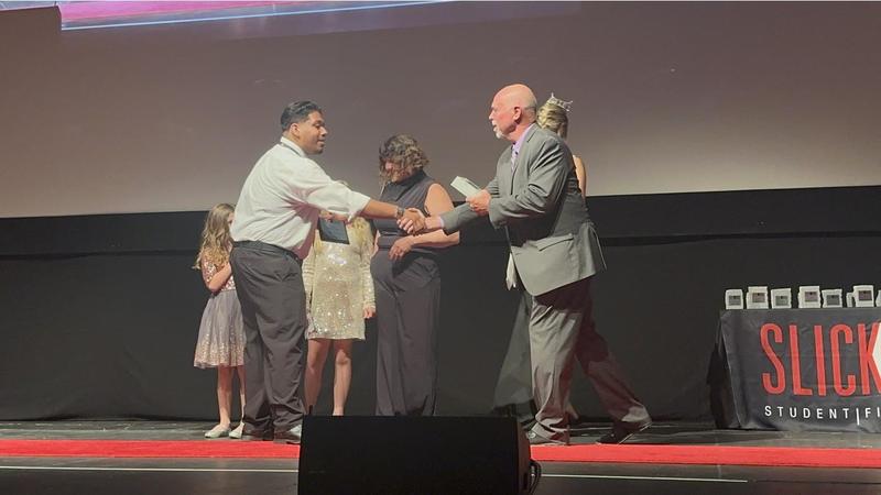 Ramses Gomez shakes hands with an award presenter on stage at the Slick Rock Student Film Festival, holding his plaque, while other presenters and children stand in the background.