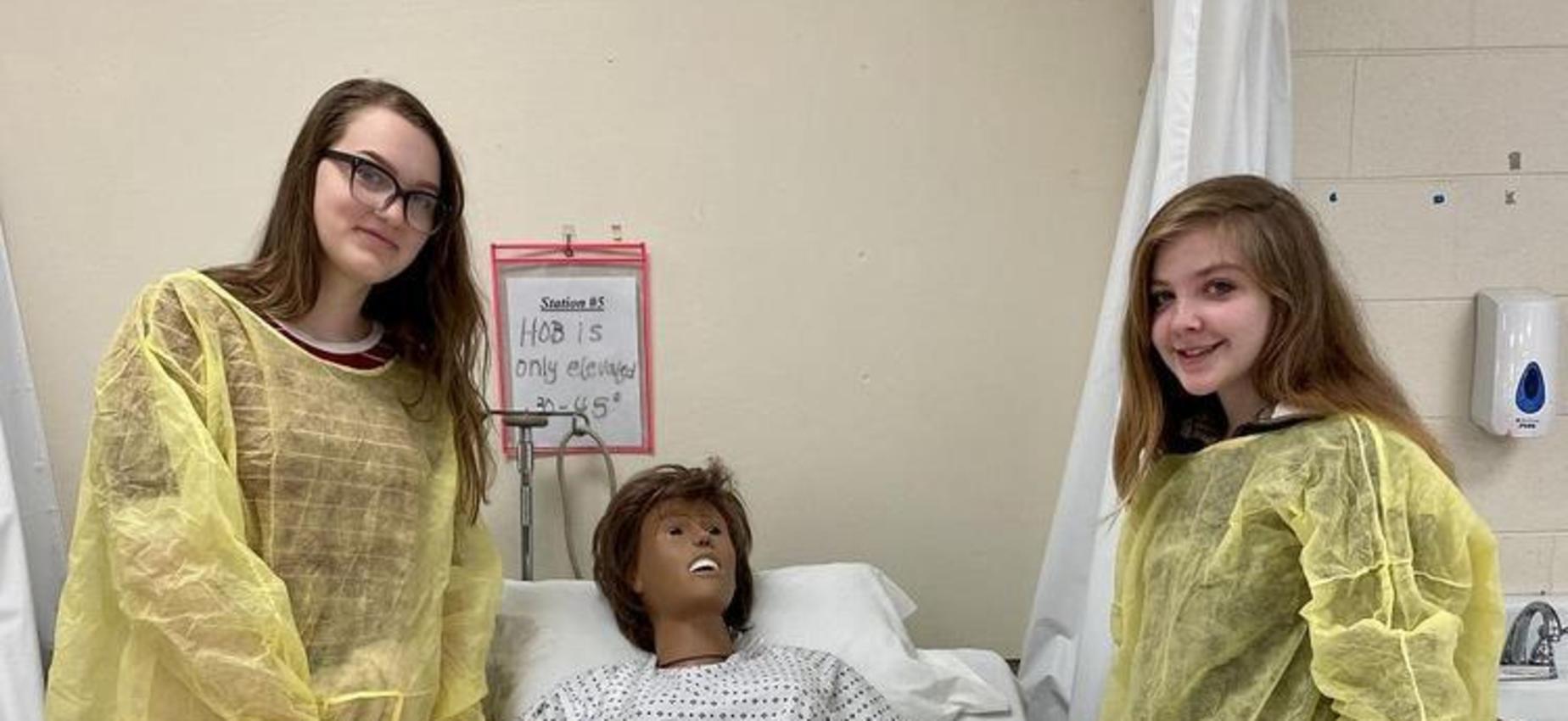 Two students stand beside a manikin in a bed.