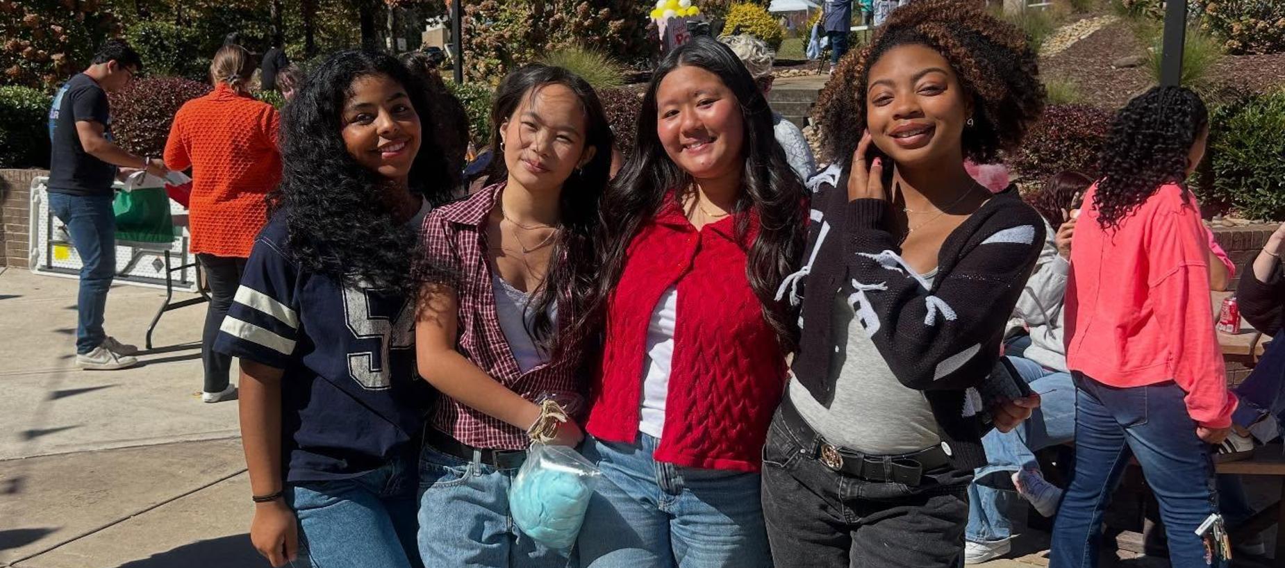Four young women posing together outdoors, smiling and wearing casual outfits.