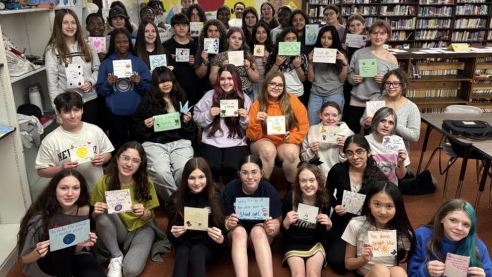 A large group of middle school students pose together in a school library, each holding up a small, hand-drawn inspirational card for Red Ribbon Week.