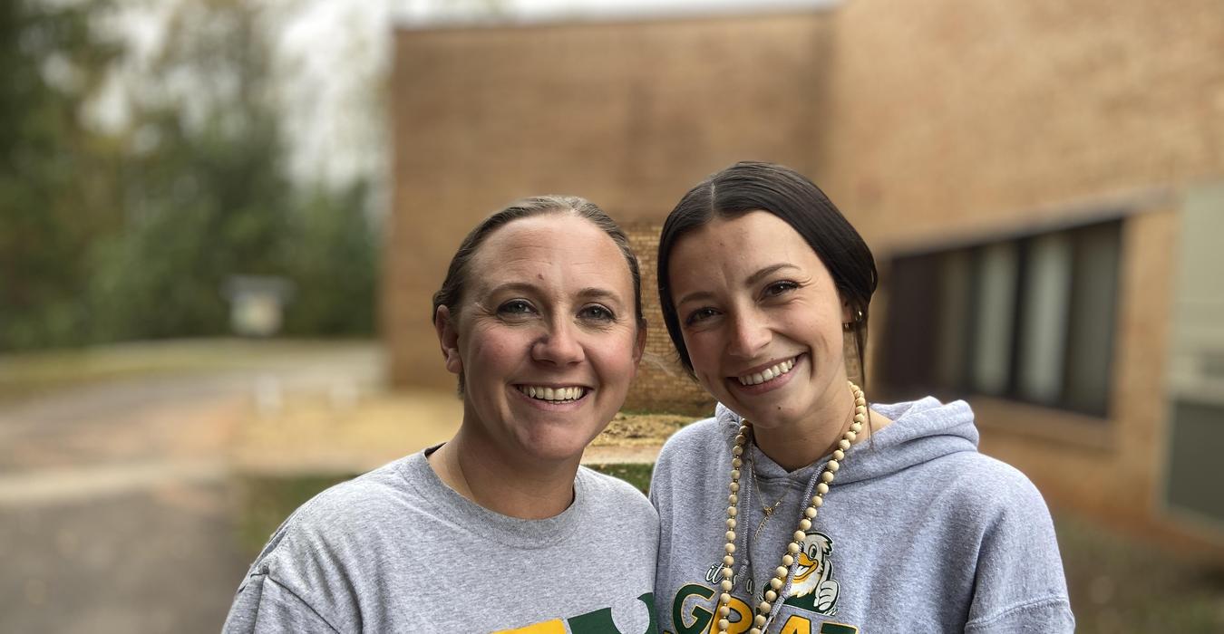 Two women smiling at the camera outdoors, wearing casual attire.