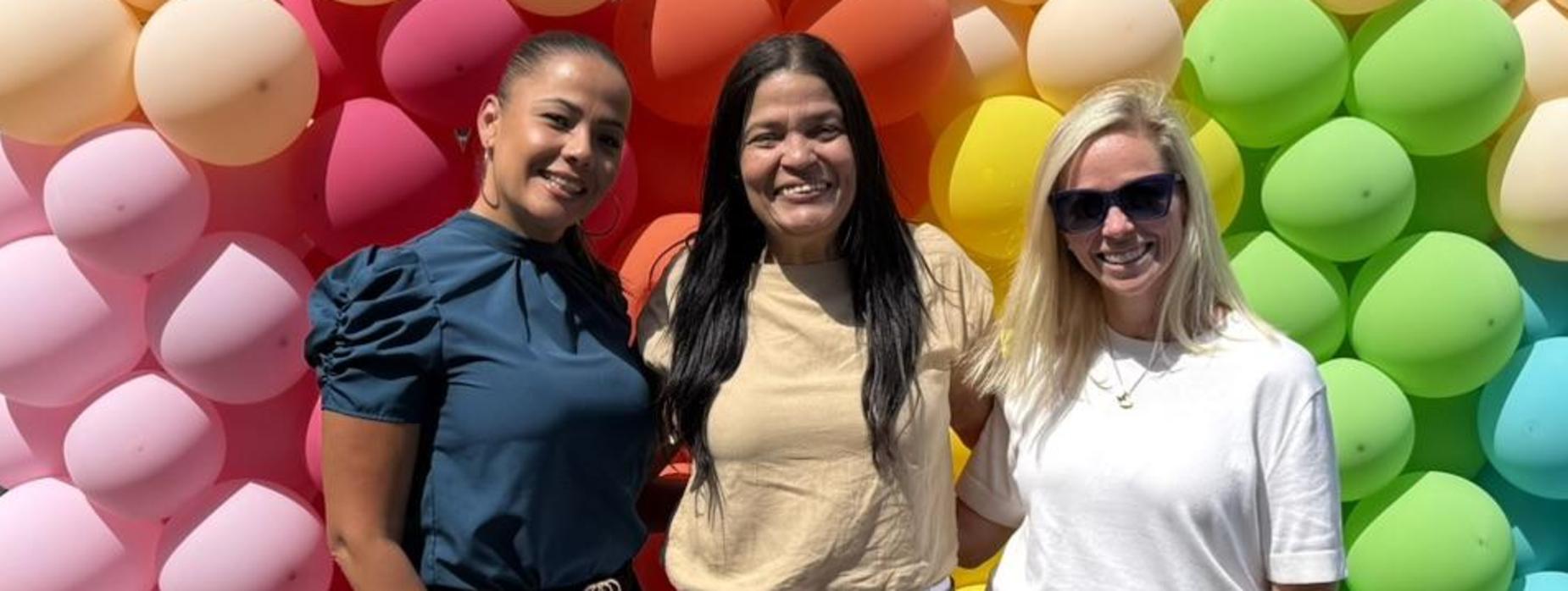 Three women pose together in front of a colorful balloon backdrop.
