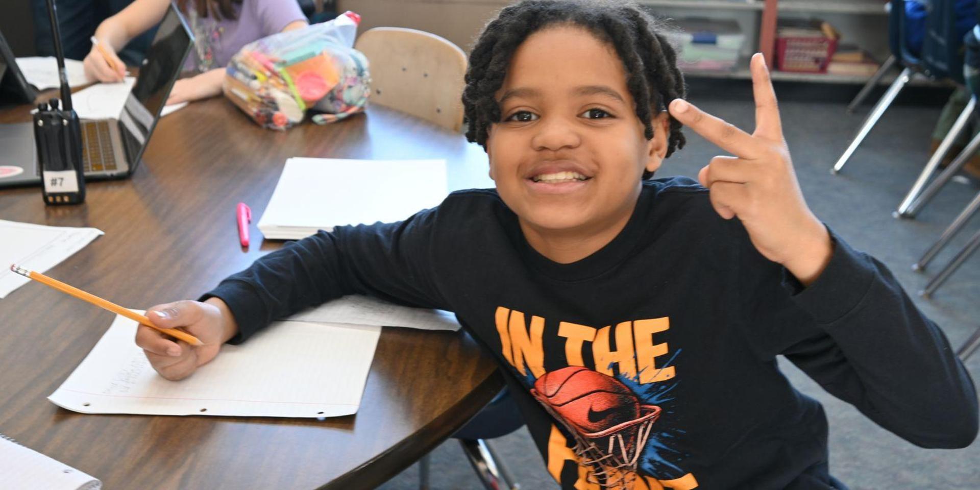 Boy showing a peace sign while working with a pencil at a desk.
