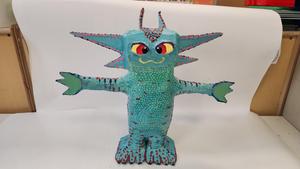 “‘Rebirth From Waste: The Dragon Axolotl,’ a student sculpture by Ahnaf Sajid. The sculpture depicts a stylized axolotl-dragon figure standing upright with outstretched arms. The body is painted turquoise blue with textured, scale-like patterns and dotted accents in pink and purple. The figure has large pointed gills extending from the head, small horn-like shapes on top, wide yellow-and-red eyes, and clawed feet. The sculpture is displayed against a plain white background.”