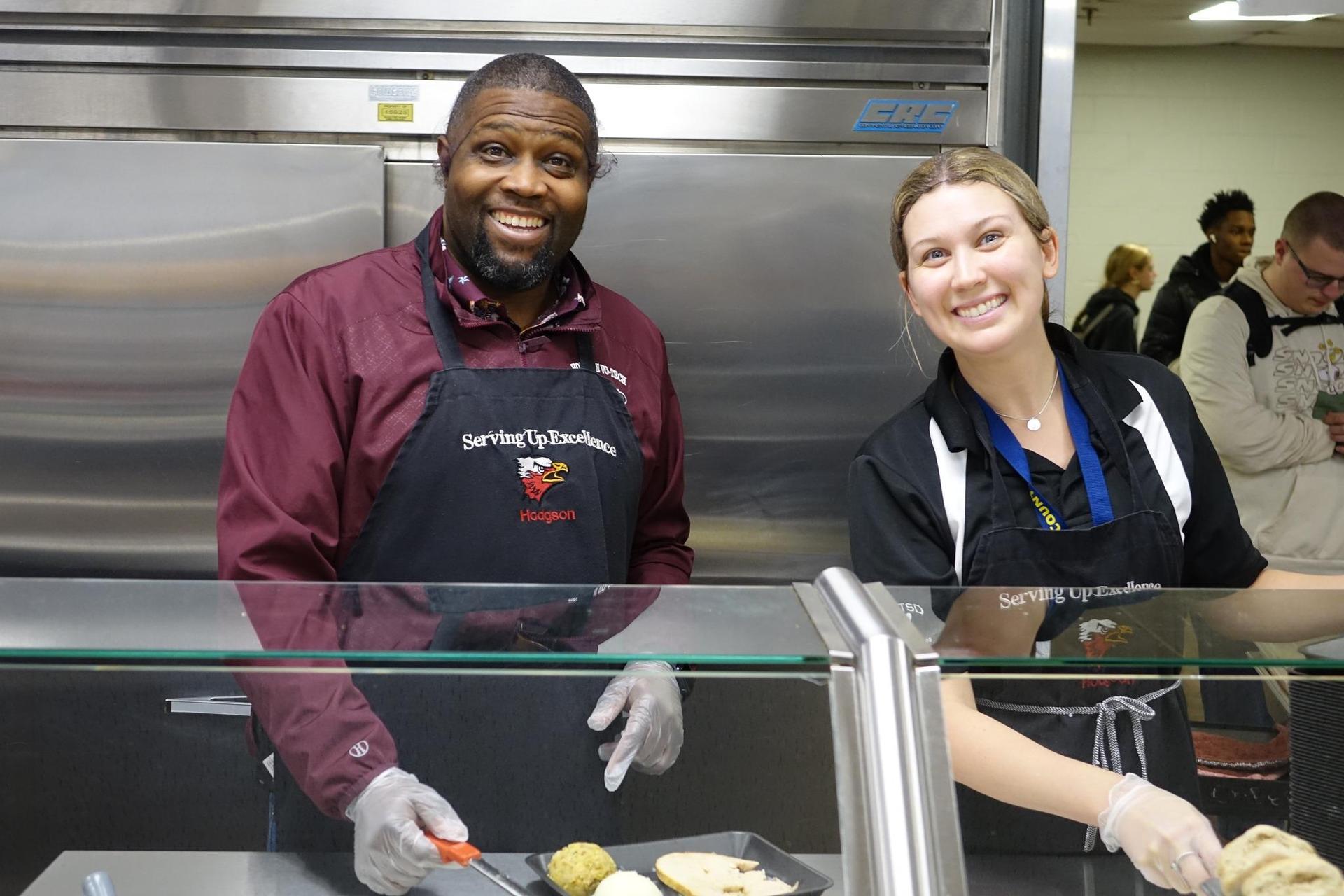 Two cafeteria staff members smiling while serving food to customers.