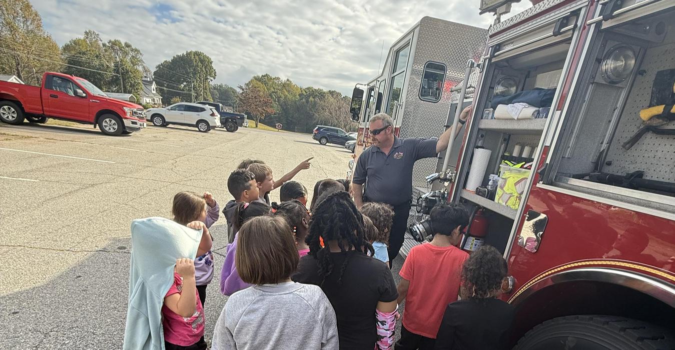 Children gathering around a fire truck, listening to an adult, in a parking lot.