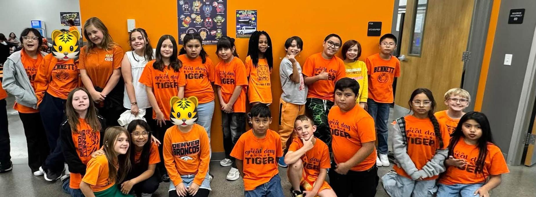 Students in orange shirts smiling and posing for a photo indoors.
