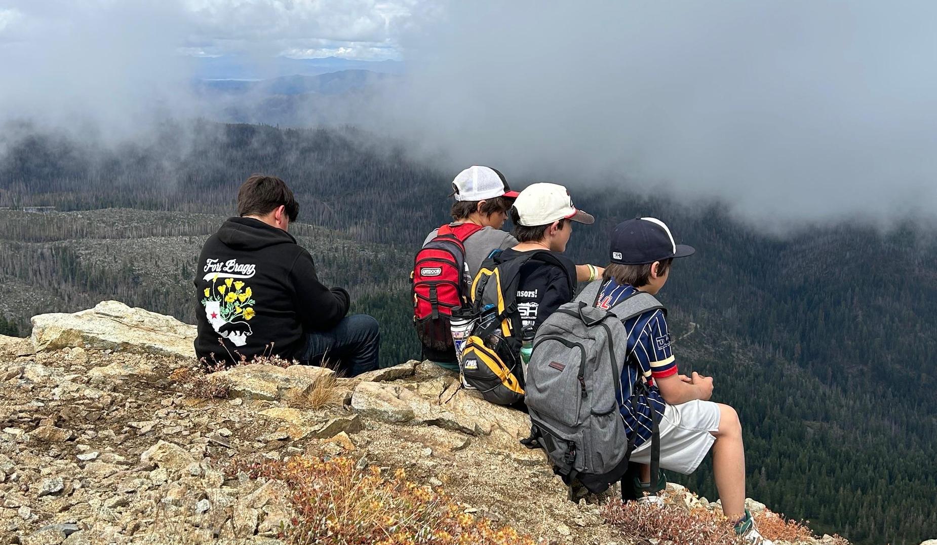 Several student perch on a mountain ledge overlooking a misty mountain forest view. They are on a hike to Spanish Peak as part of an outdoor education trip.