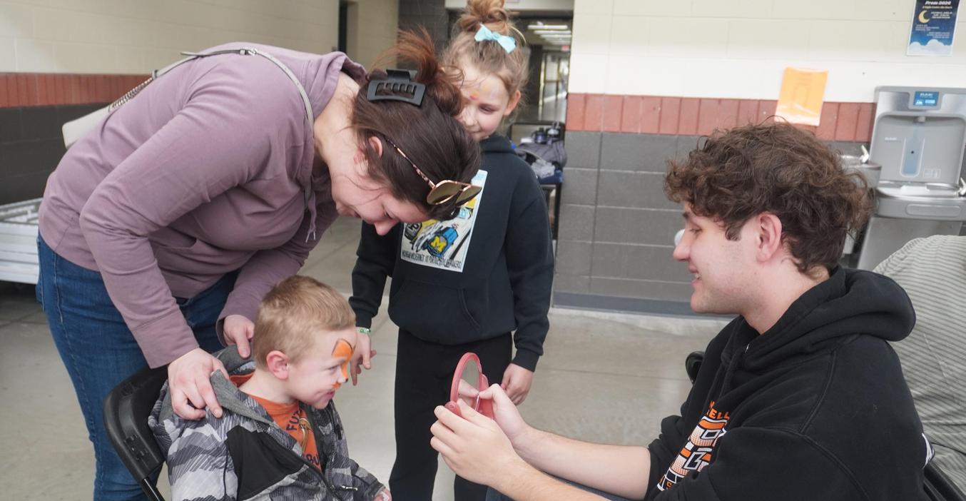 A high school student holds up a mirror for a young boy to see the face painting he did.