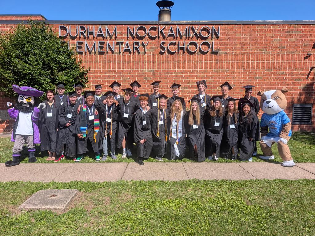 Group photo of senior graduates in caps and gowns standing in front of the building sign.