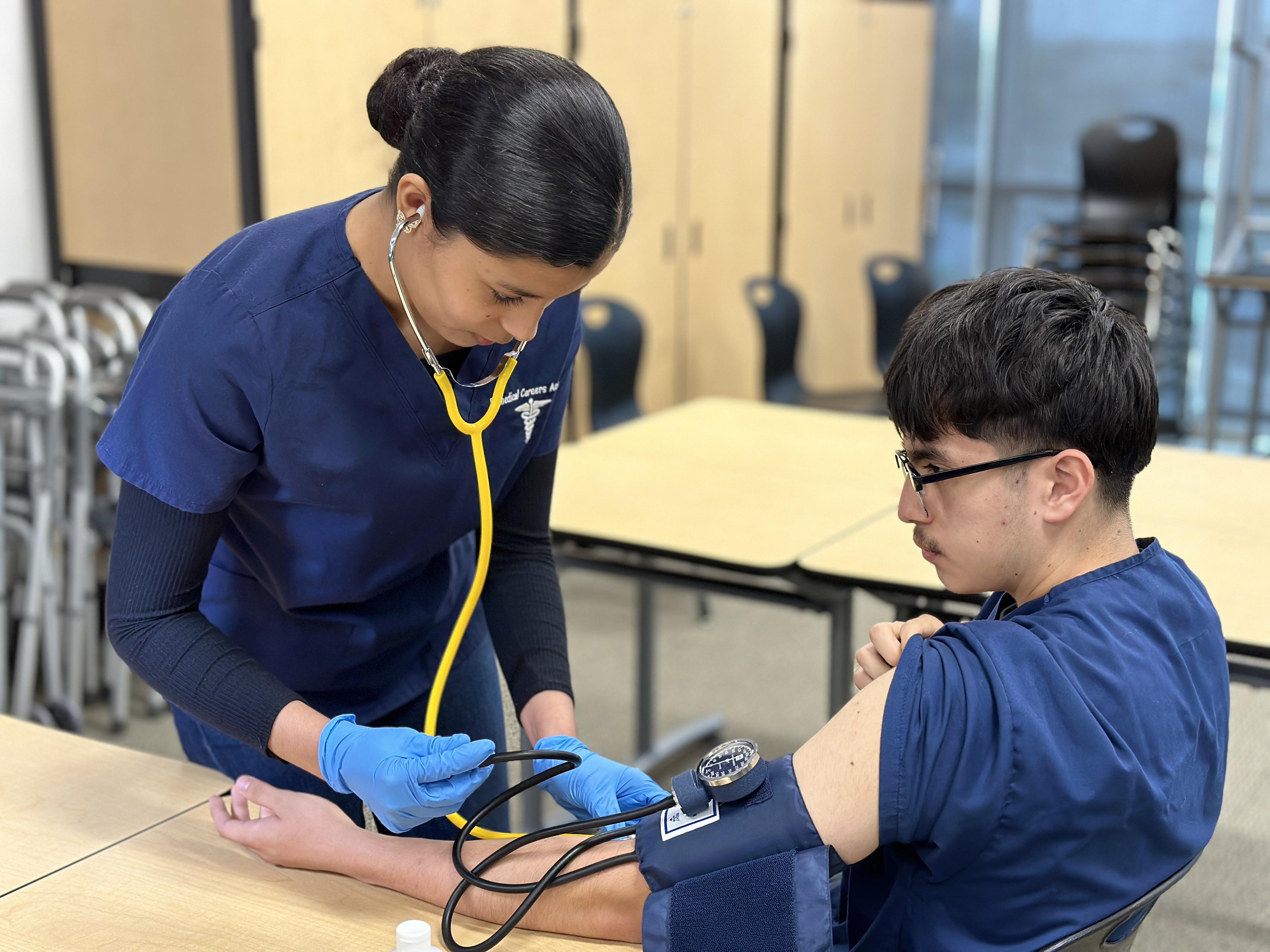 Two students in medical scrubs practice taking blood pressure in a classroom, with one student using a stethoscope and blood pressure cuff on the other student’s arm.