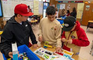 Three students collaborate on a hands-on engineering activity, selecting parts from a LEGO Education kit at a classroom table. The room is filled with colorful supplies, posters, and active student engagement in the background.
