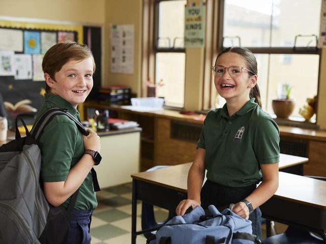 Two smiling schoolchildren with backpacks in a classroom setting.