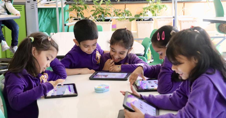 A group of five children in purple shirts using tablets at a table in a classroom.