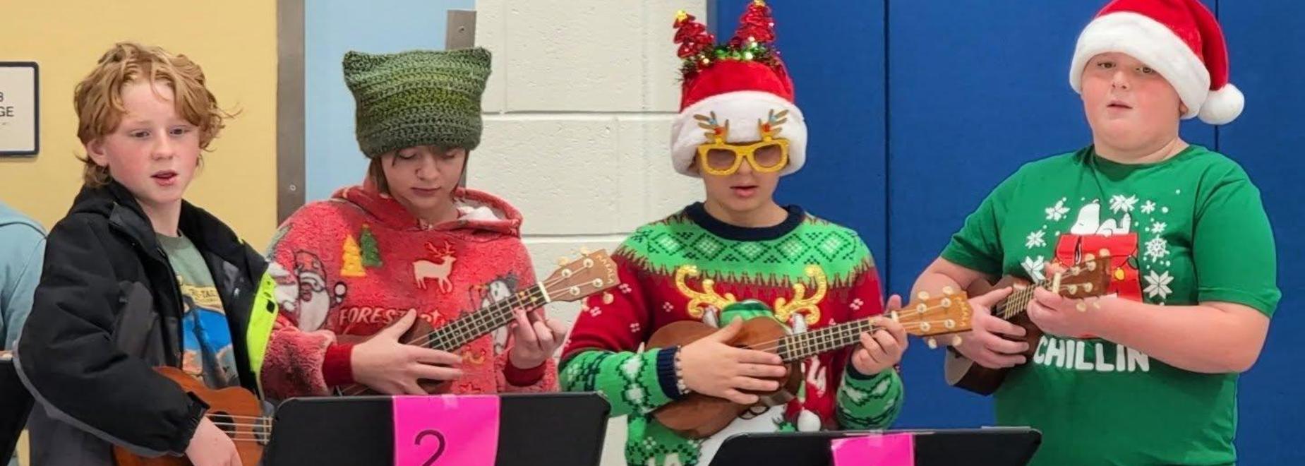 Four children in holiday sweaters play ukuleles while wearing winter hats and Santa hats.