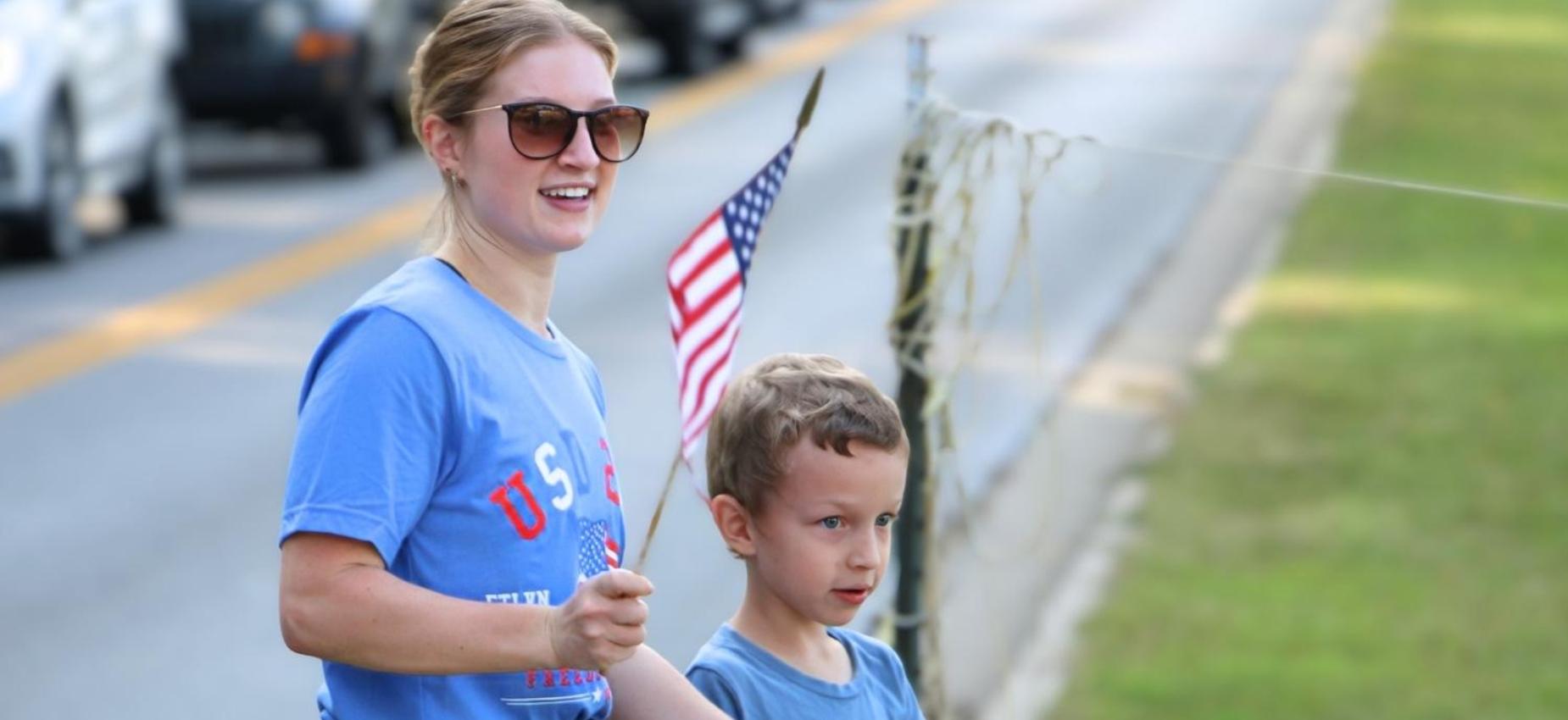 Woman and child waving an American flag beside a road.