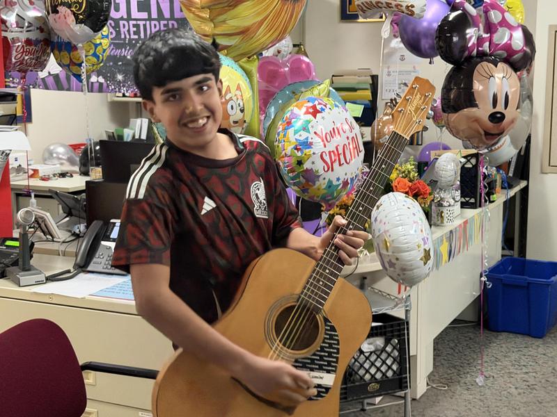 Boy holding an acoustic guitar with colorful balloons and decorations in the background.