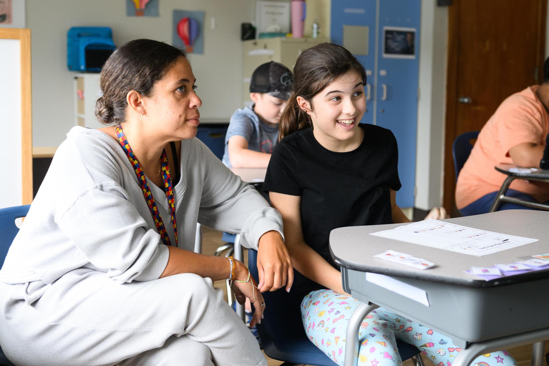 A student sitting at the desk smiling, teacher is sitting next to her.