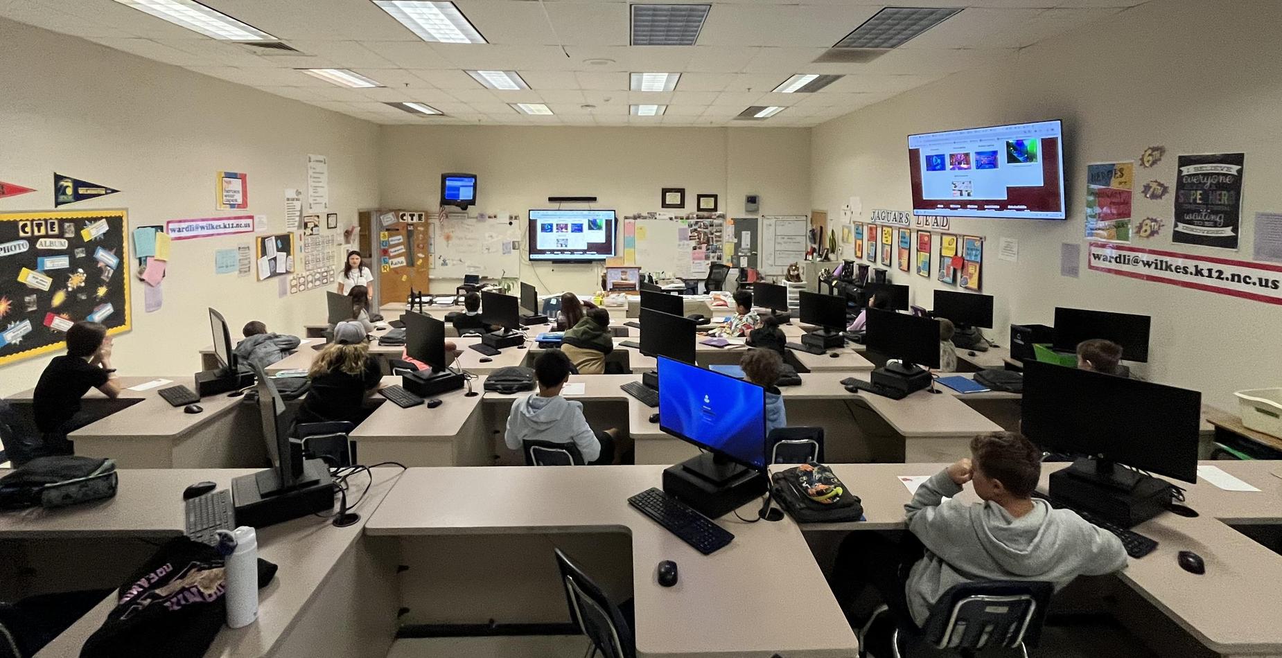 Interior of a classroom with students at computers and a teacher at the front.