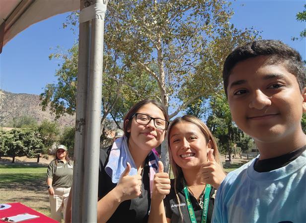 Three smiling individuals giving thumbs up; one person stands in background outdoors.