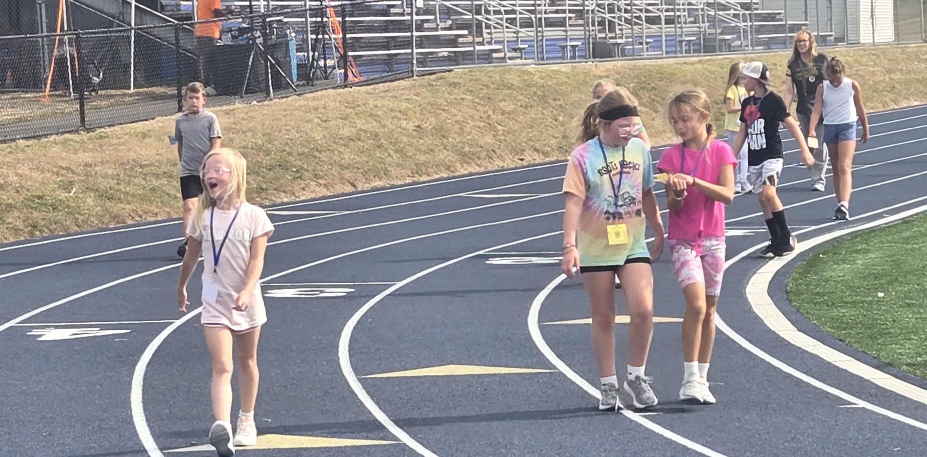 Two girls chatting and walking on the track, with other children in the background.