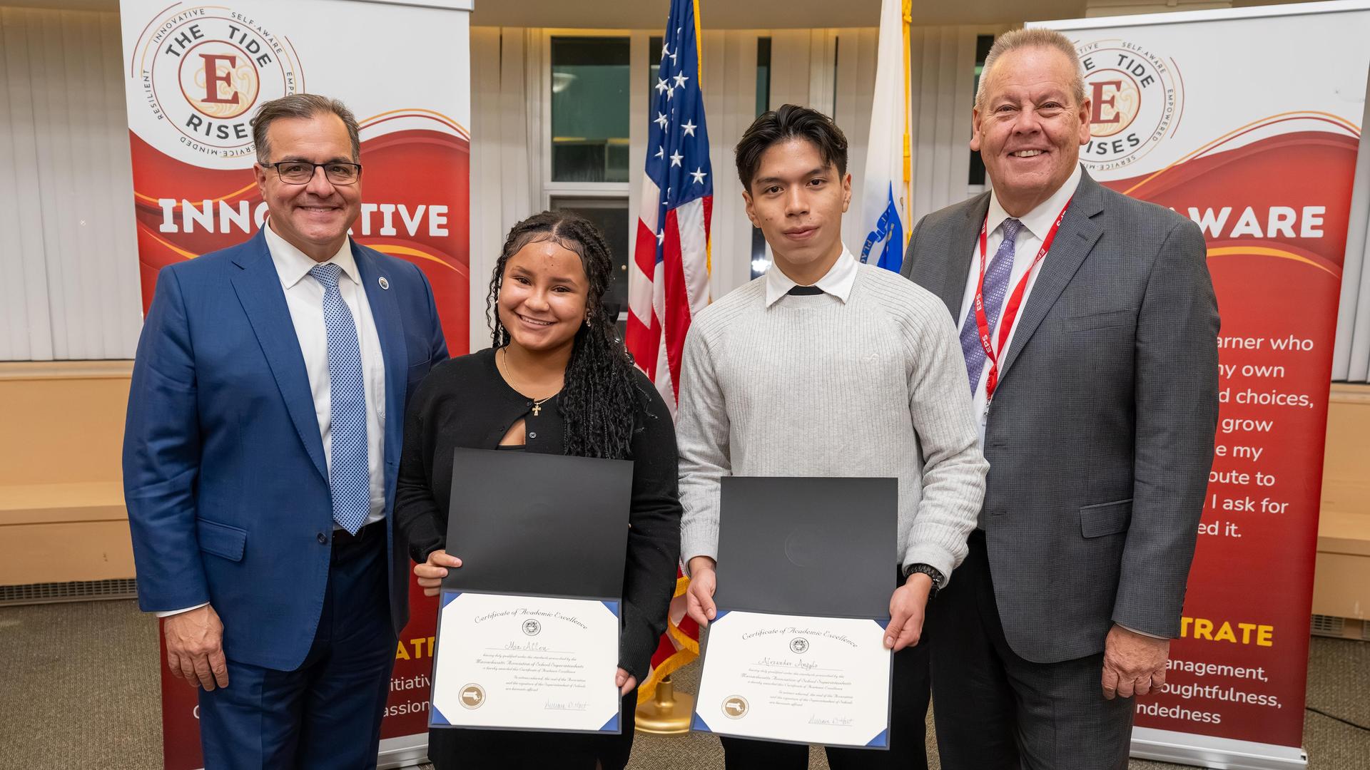 Two students holding certificates stand with officials in front of flags.