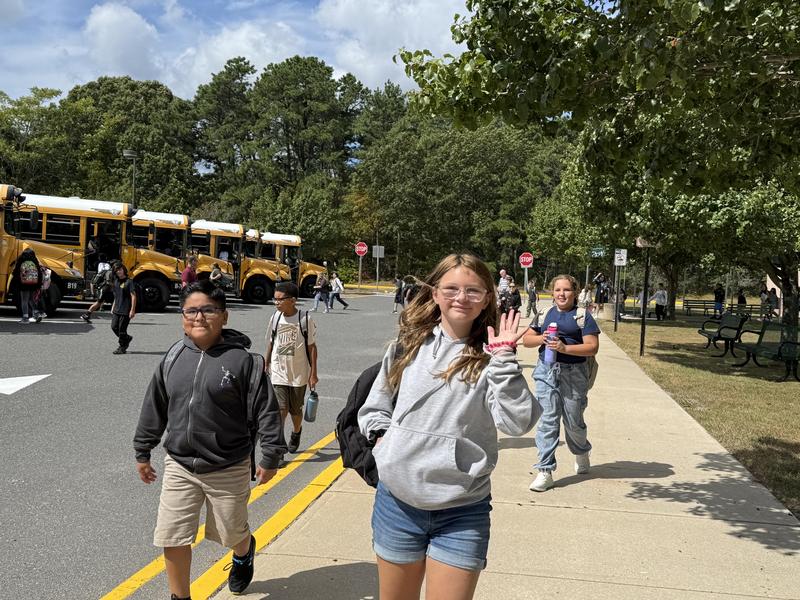 Students walking on a school sidewalk, smiling and waving near yellow school buses.