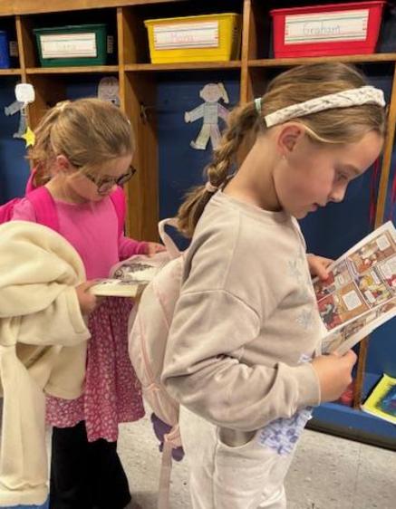 Two girls in a classroom, one reading a book while the other carries a backpack.