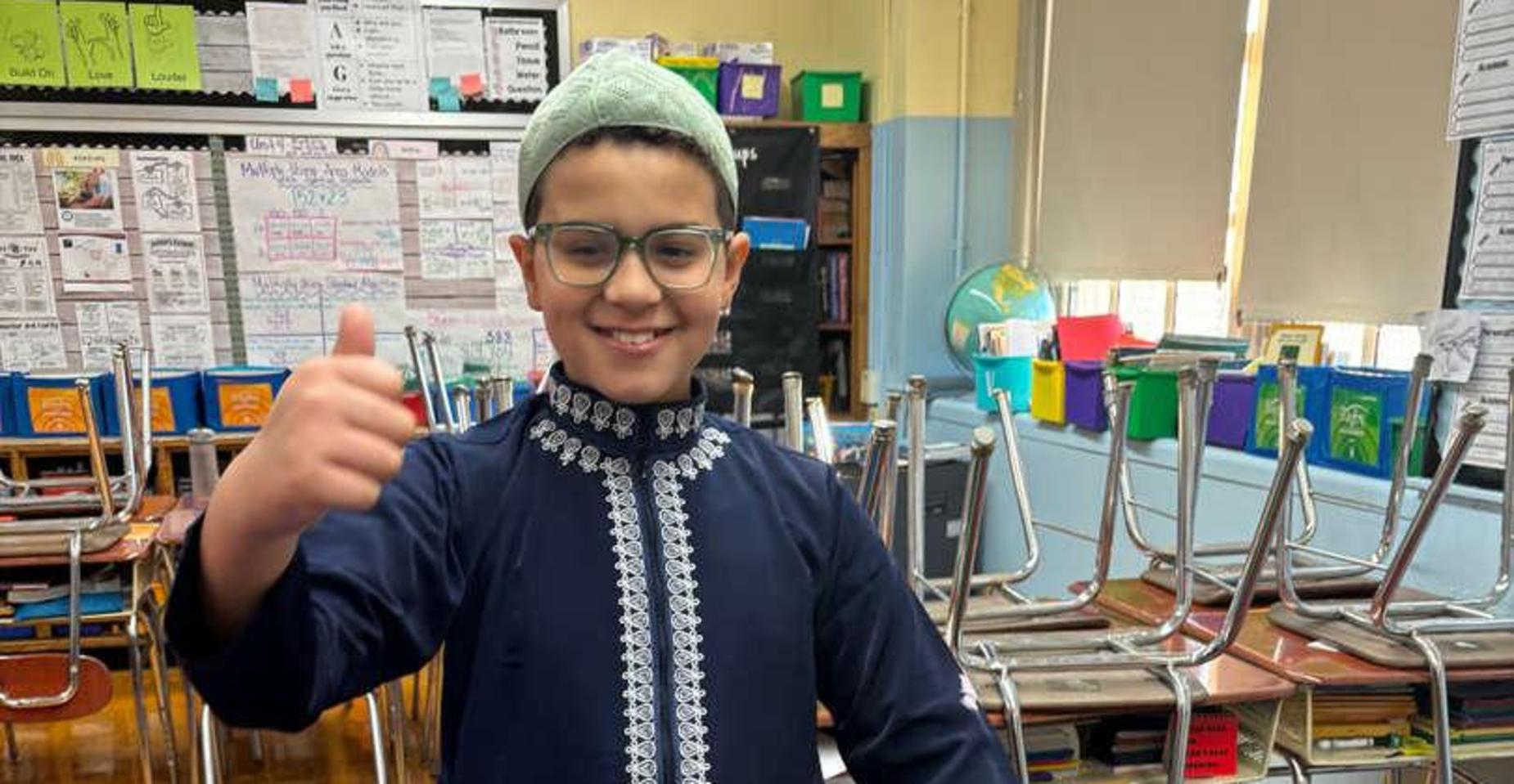 Child in traditional attire smiling and giving a thumbs up in a classroom.