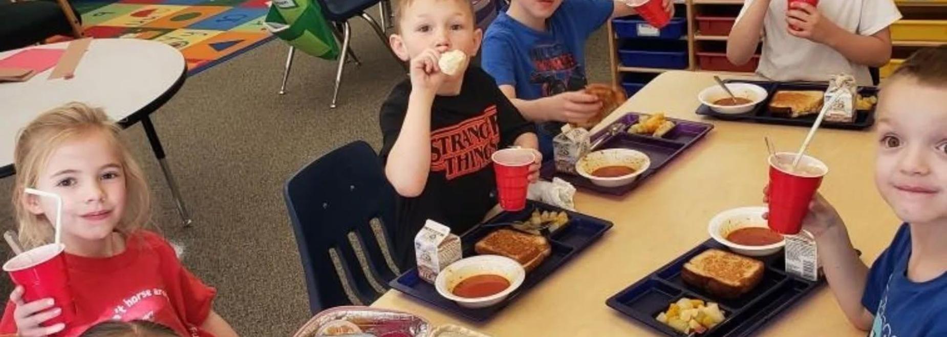 Group of children enjoying lunch with trays and drinks in a classroom.