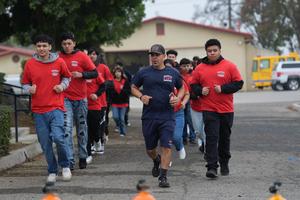 A group of students in red Porterville Fire Academy shirts jog together outdoors, led by an instructor in navy athletic gear. The scene captures a team-building fitness drill on a school campus.