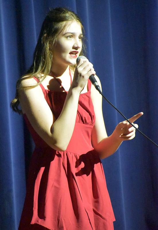 A singer in a red dress gestures while singing on stage.