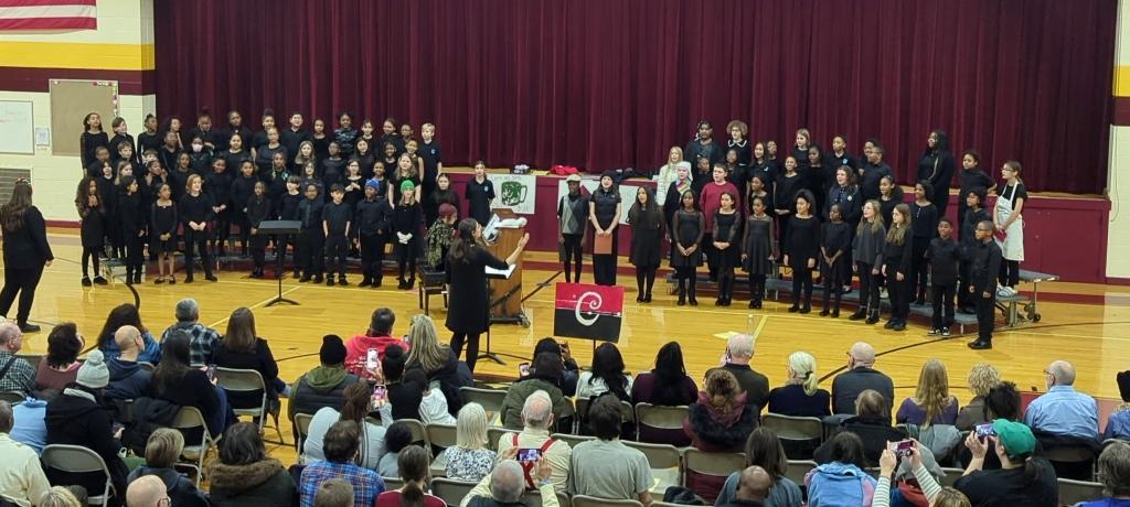 teacher directs large choir of singing children standing on risers in the gym