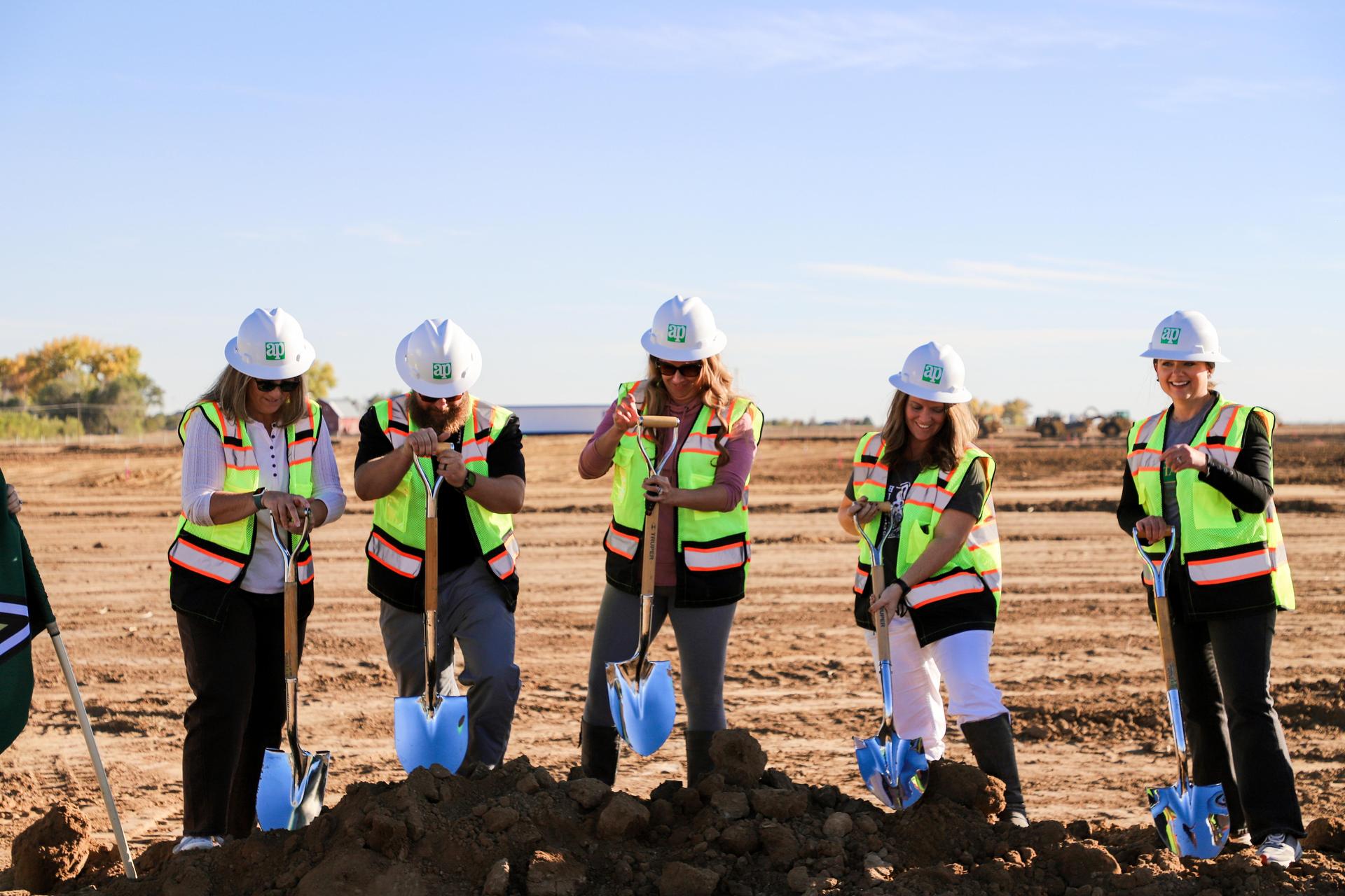 Five women in hard hats and vests posing with shovels at a construction site.