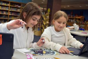 Two girls smile while playing a game with tablets in a library.