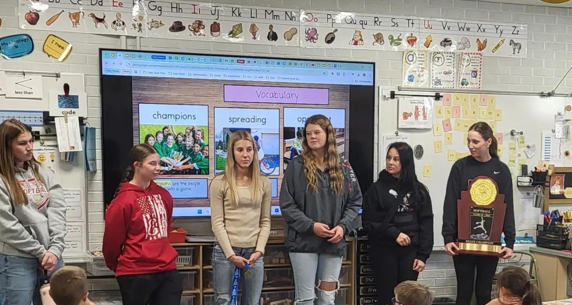 Group of students presenting in a classroom with a trophy and young kids watching.