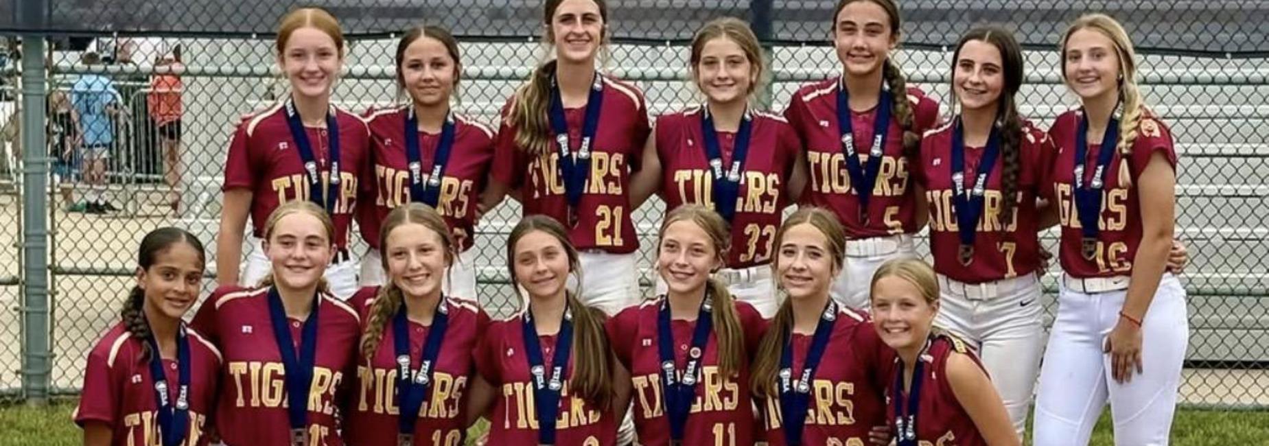 A girls' softball team poses together holding a trophy, wearing medals and uniforms.