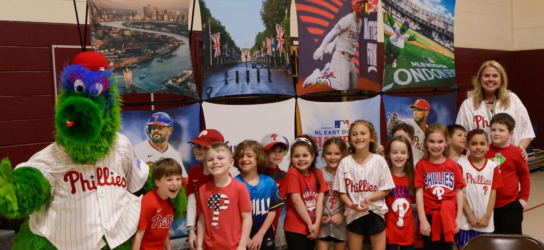 A group of children in Phillies gear with the Phillie Phanatic and their teacher.