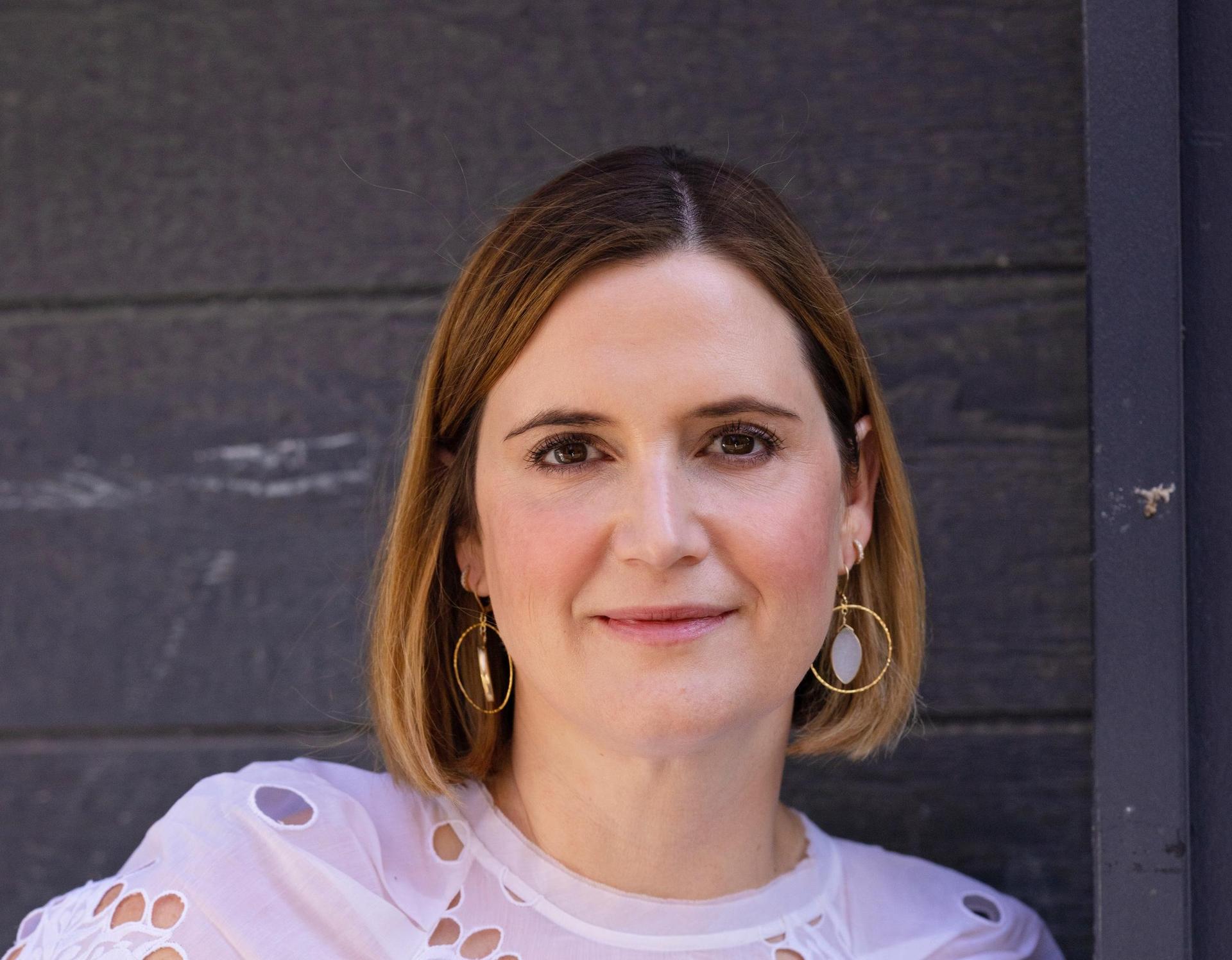 A woman with short brown hair, wearing gold hoop earrings and a white blouse, smiling at the camera.