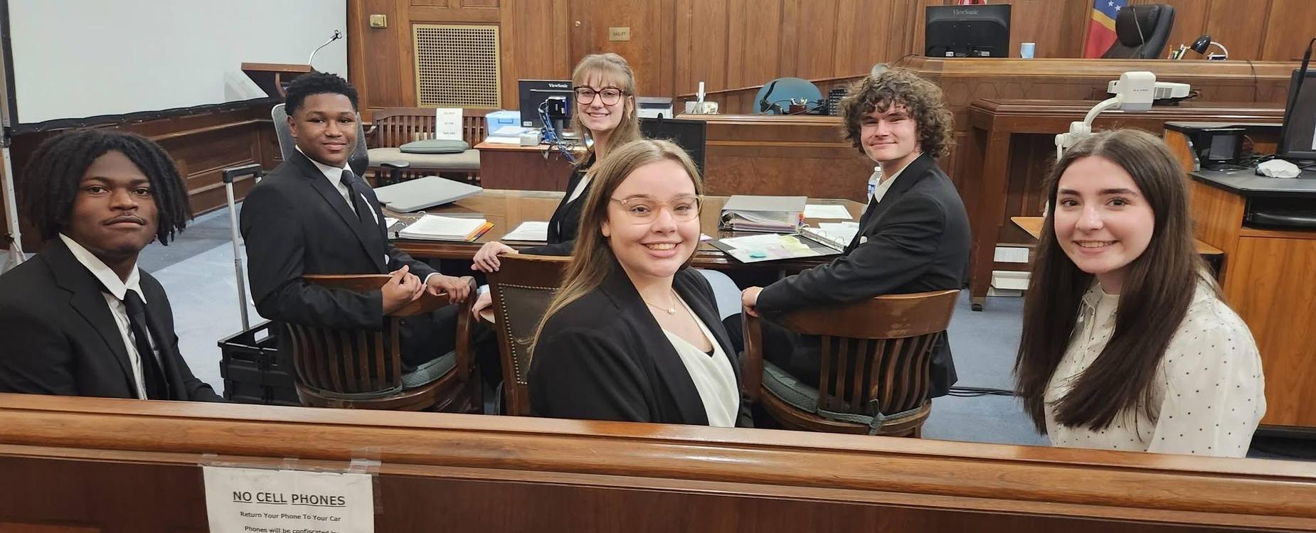 Group of five people in a courtroom setting, seated and smiling at the camera.