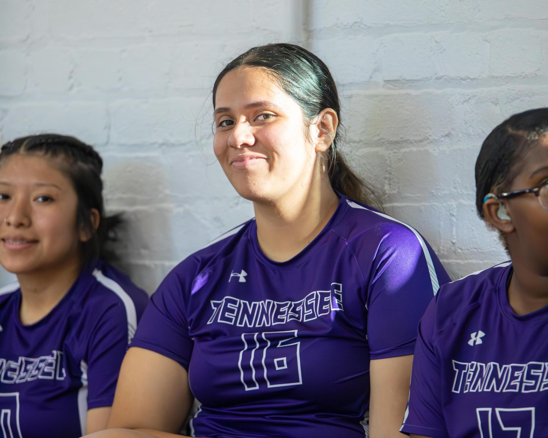 A smiling volleyball player in a purple jersey sits with teammates in the background.
