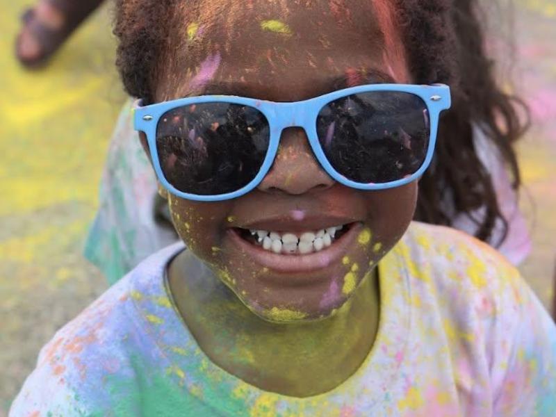 A girl with sunglasses smiles brightly, covered in colorful powder.