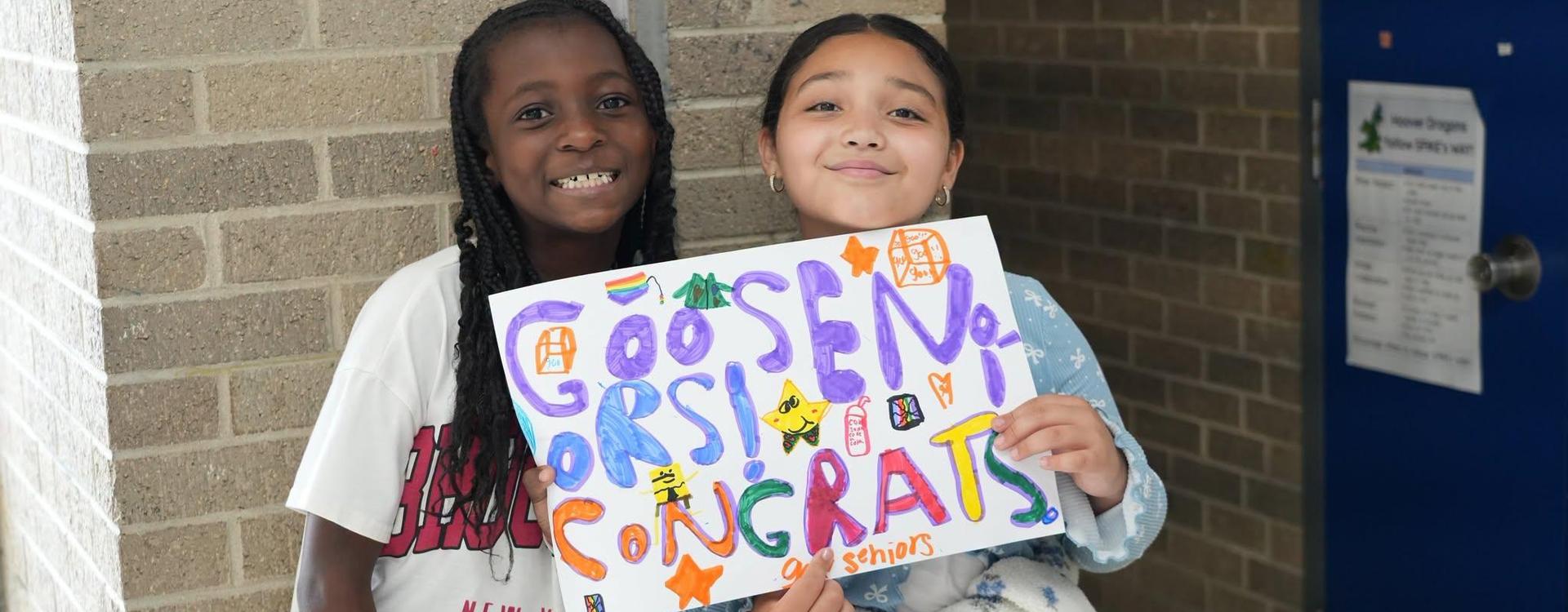 Two girls smile while holding a colorful congratulatory sign for seniors.