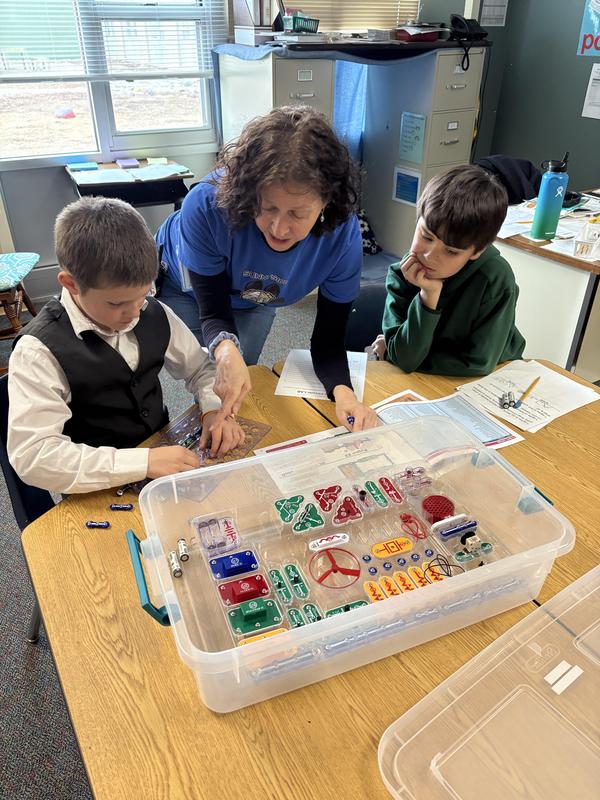Teacher assisting two children with a science activity at a classroom table.
