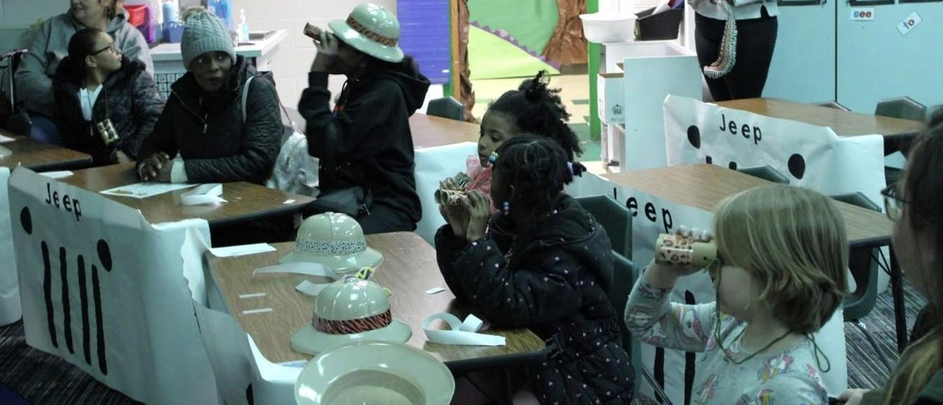 Students wear hats and look through toy binoculars at safari-themed desks in a classroom.