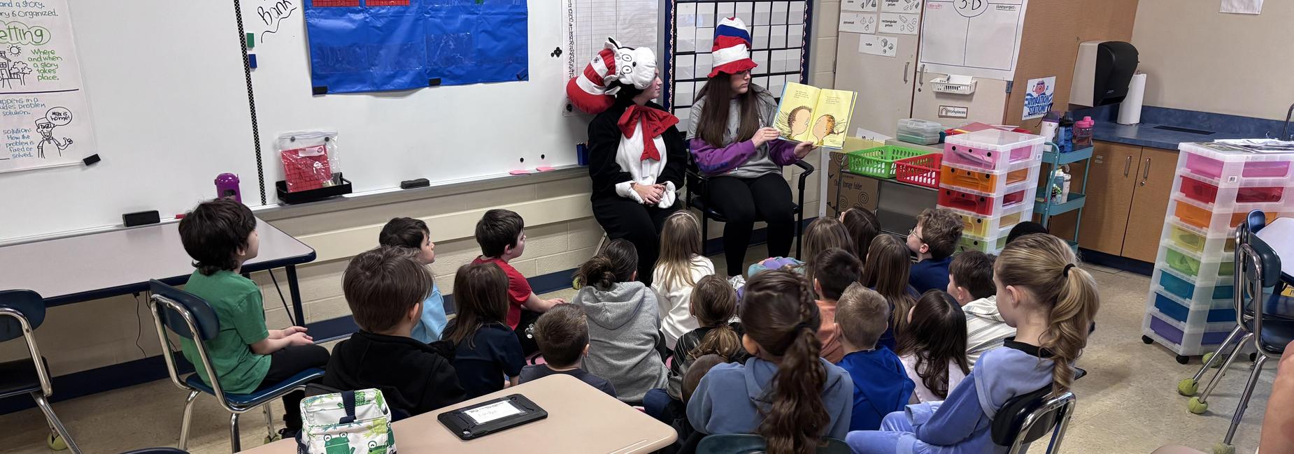 Students in chairs listening to guest speaker reading a book.
